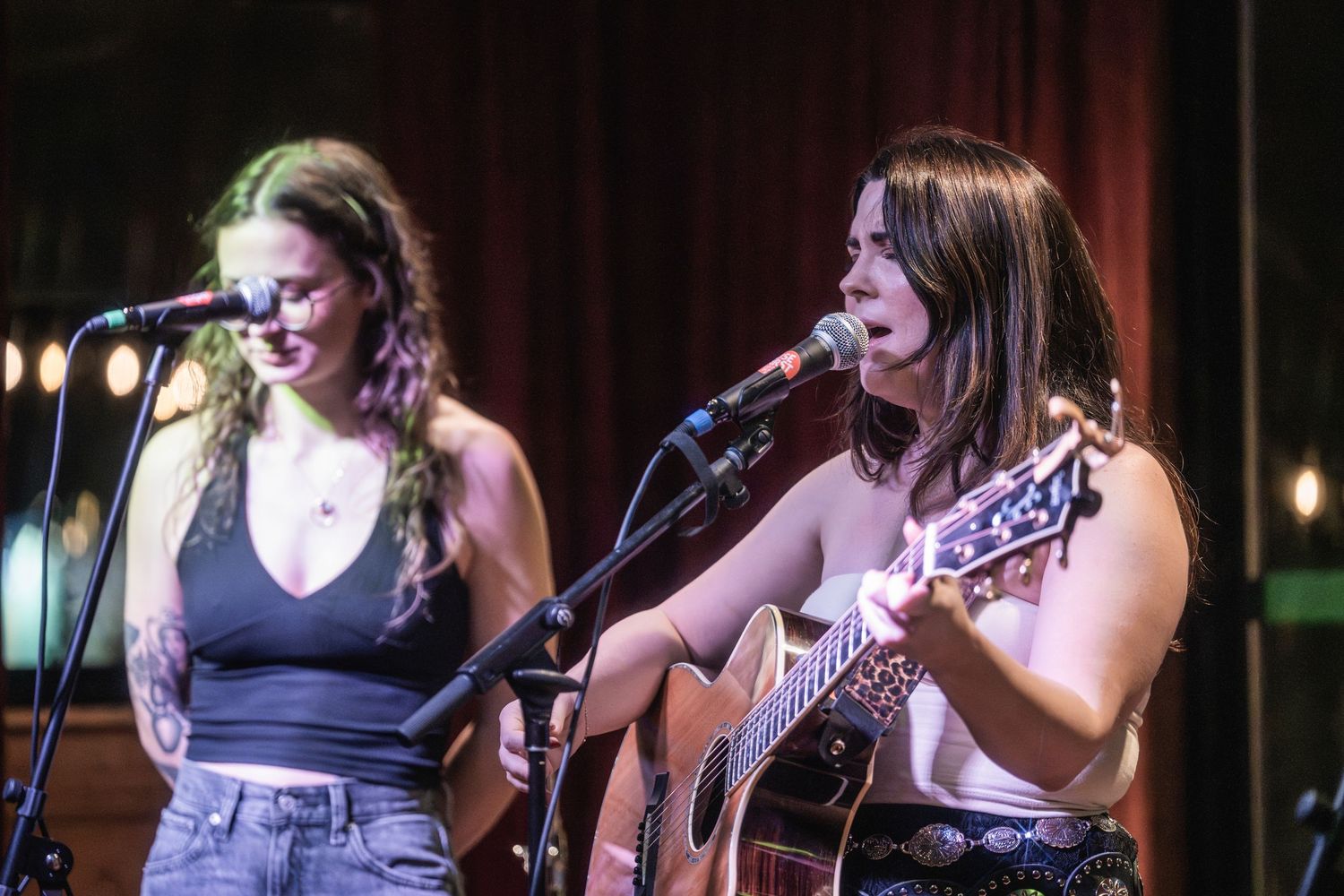 Two musicians perform on stage, one singing into a microphone while the other plays acoustic guitar under warm lighting.