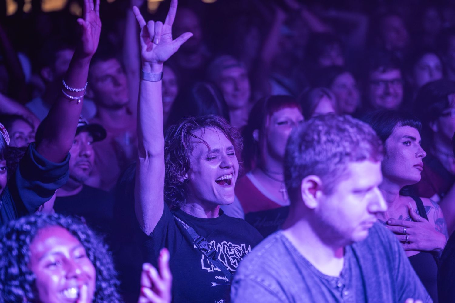 Concert audience members smile and cheer while bathed in purple stage lighting at a live music performance.