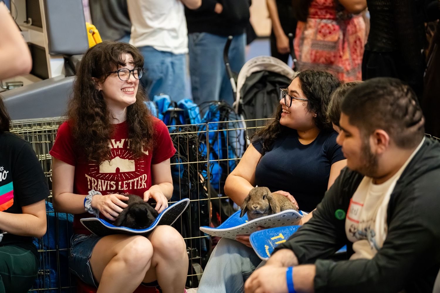 Students smile and interact while holding small puppies at an indoor event, with cages and backpacks visible nearby.