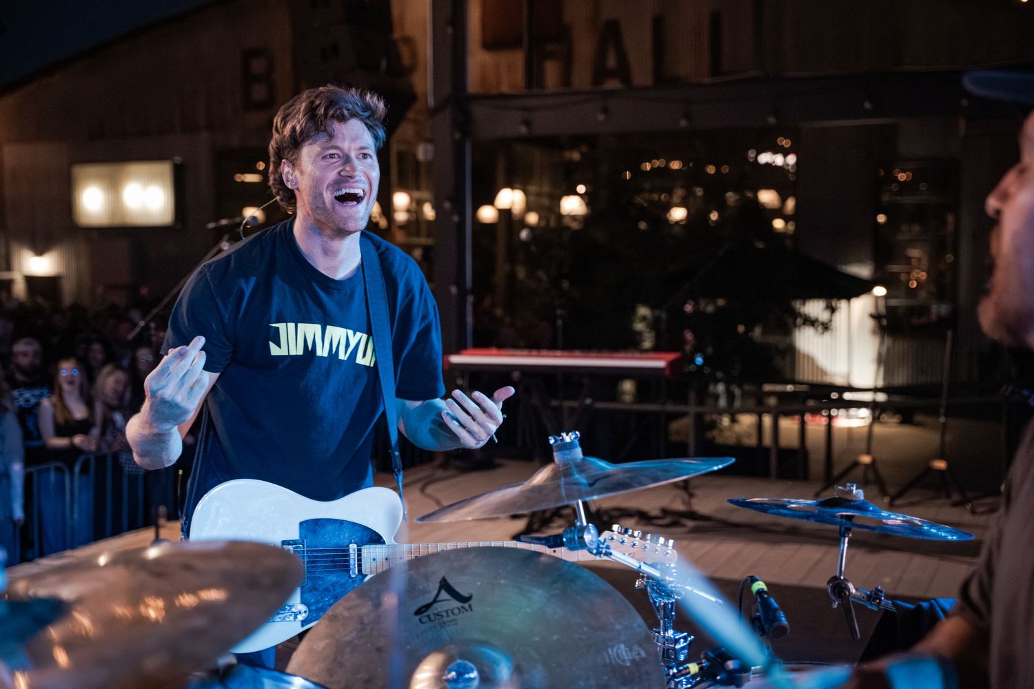 A male musician performs energetically on stage playing guitar, wearing a black hoodie, with drums and cymbals visible nearby.