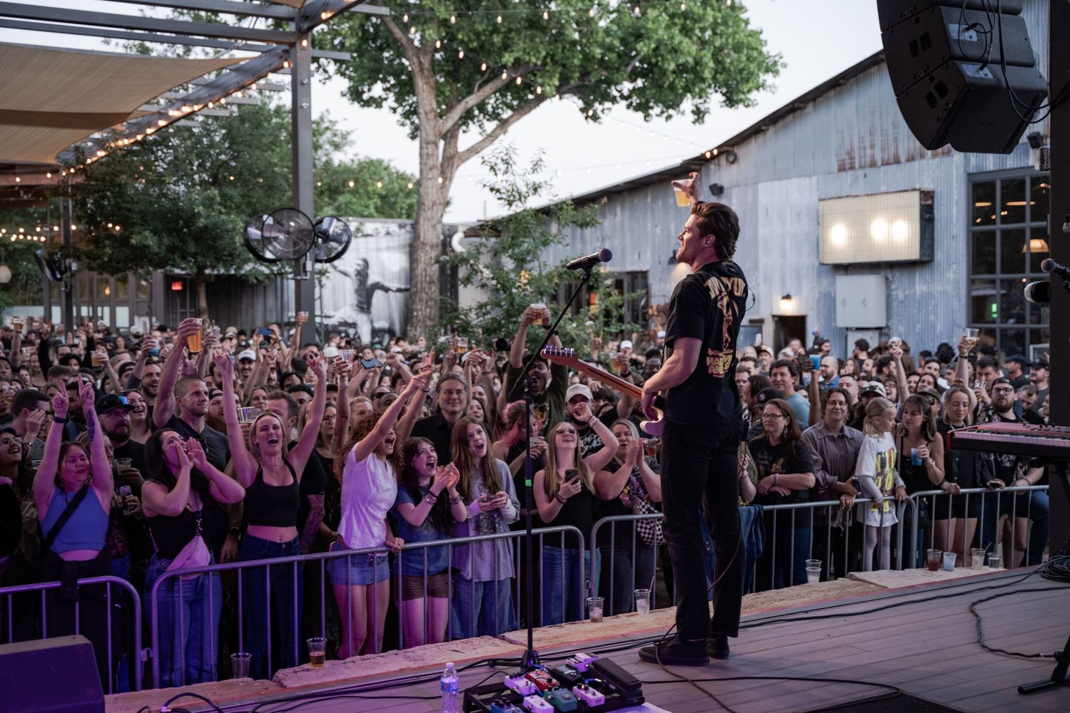 A performer on stage faces an enthusiastic crowd at an outdoor concert venue at dusk.
