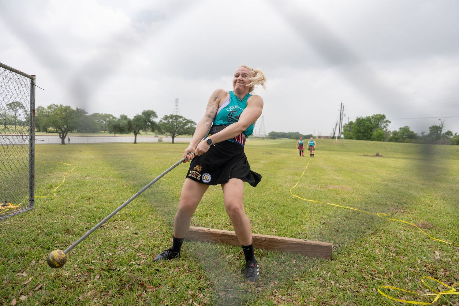 Athlete in teal top and kilt competes in hammer throw at Highland Games on grassy field.