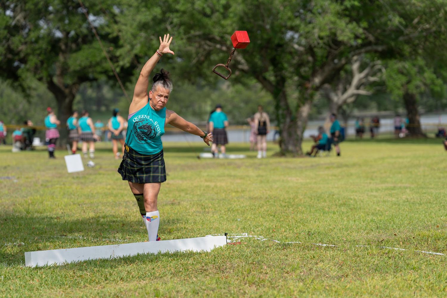 Athlete in kilt throws red object at Highland Games competition on green field with trees.