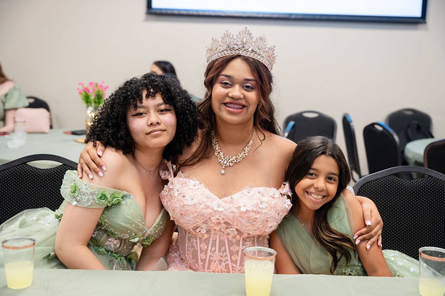 Three girls in formal dresses pose together at a quinceañera celebration, with the birthday girl wearing a pink gown and tiara.