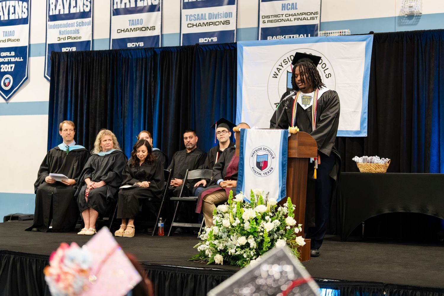 Graduate delivers speech at podium during commencement ceremony with faculty seated on stage in decorated gymnasium.