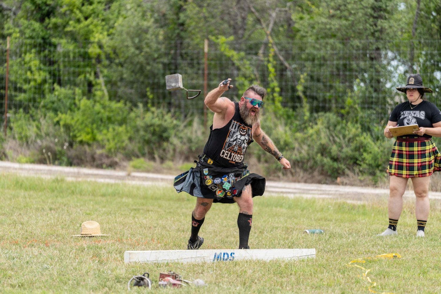 Man in kilt throws hammer at Highland Games competition outdoors, with spectator in tartan watching nearby.