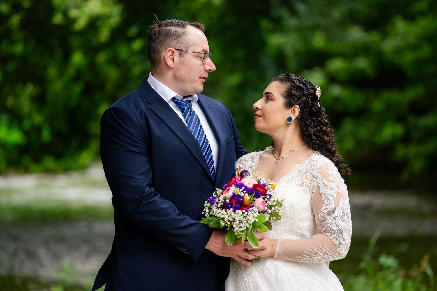 Bride and groom share a romantic moment outdoors, bride holding a vibrant purple and pink bouquet, both in formal wedding attire.