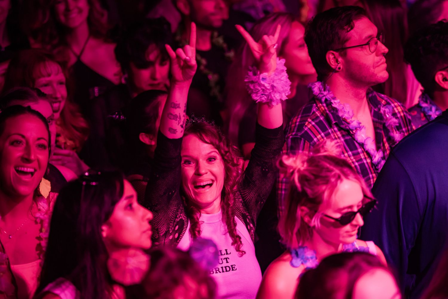 Excited concert crowd bathed in pink light, fans cheering and making rock and peace signs at a live music event.