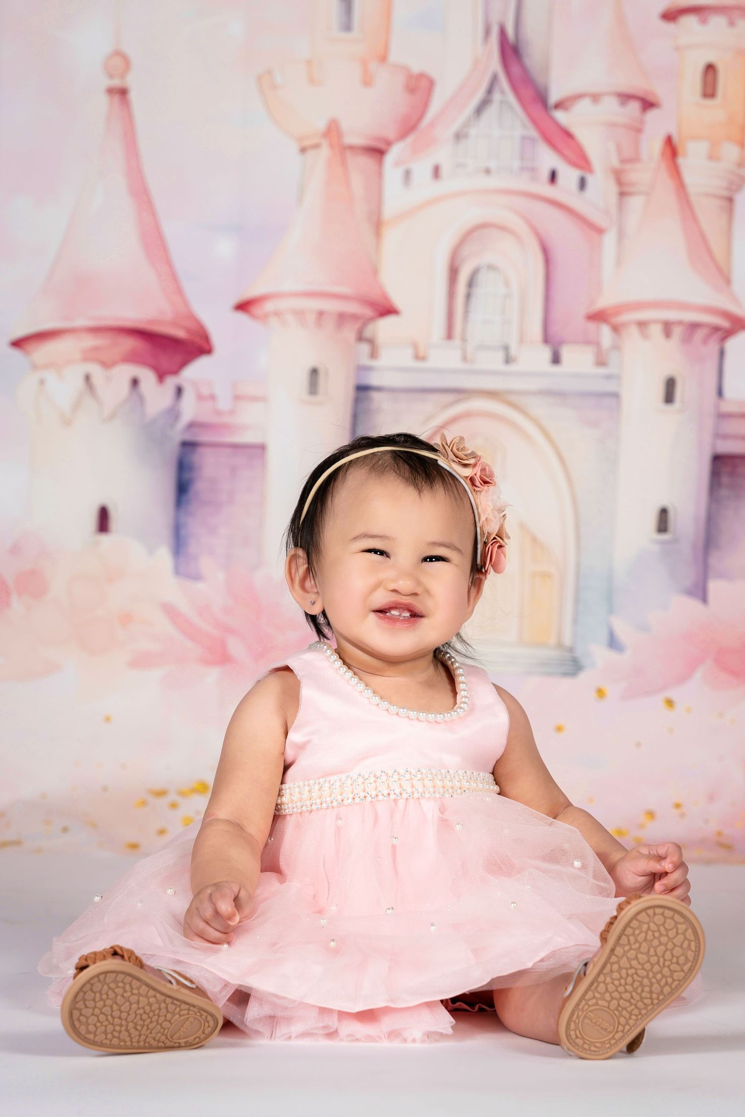 A smiling baby girl in a pink tulle dress sits in front of a watercolor princess castle backdrop.