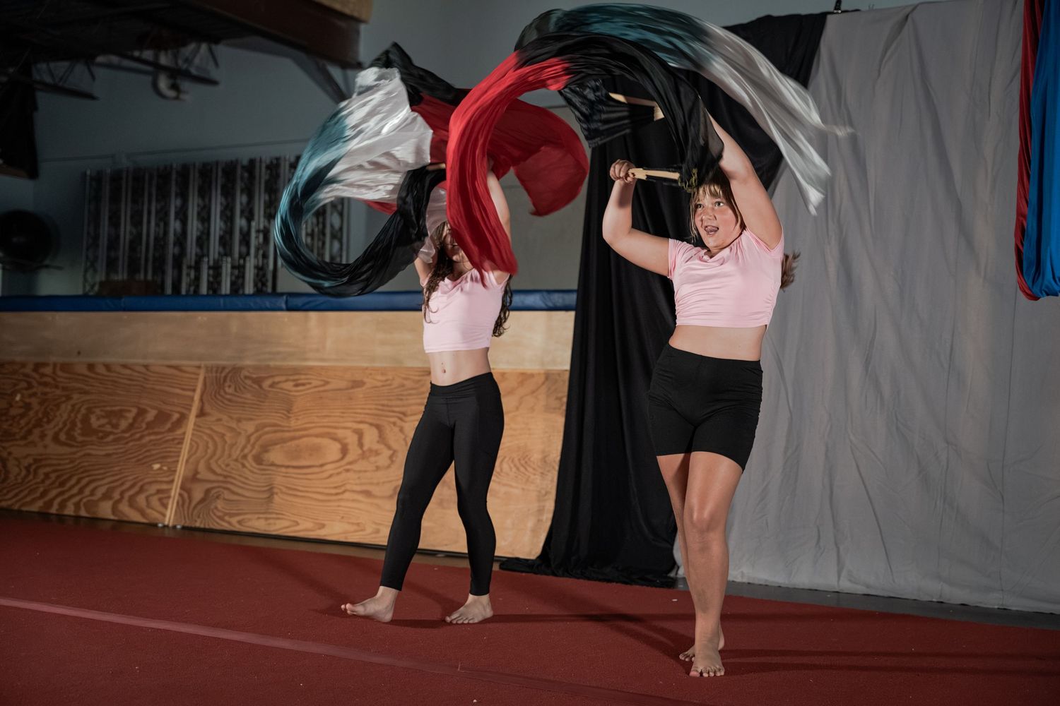 Two women performing flag dance routines indoors, waving colorful red, white, and black flags in a gym.