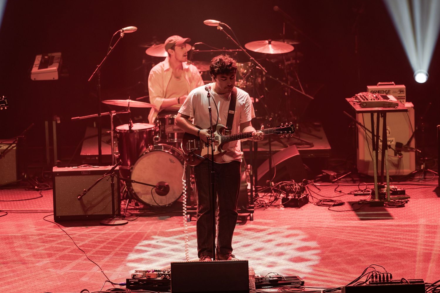 Two musicians perform on stage, one playing guitar and one on drums, bathed in red concert lighting.