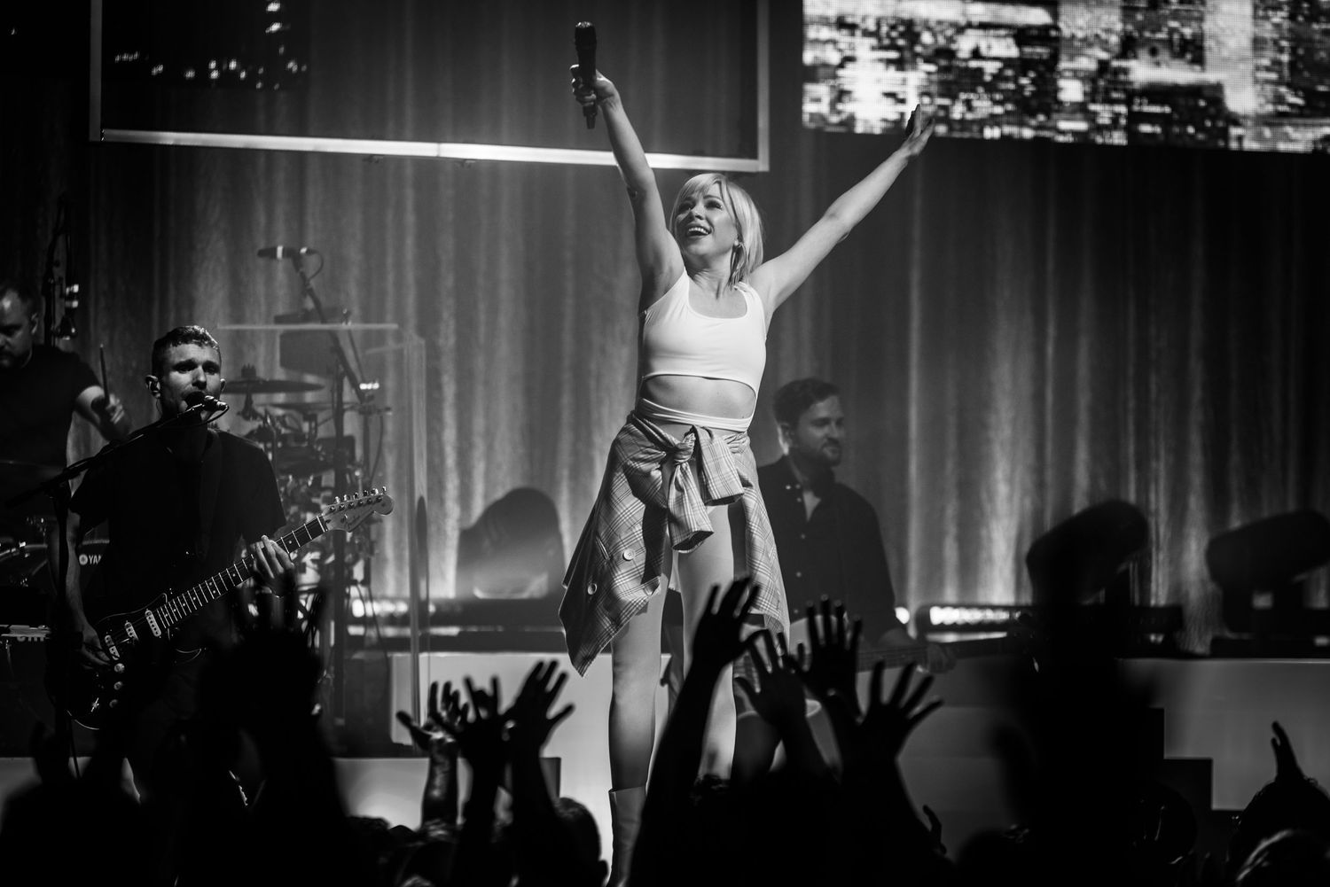 A performer stands with arms raised triumphantly on stage during a concert with silhouetted crowd and band members visible.