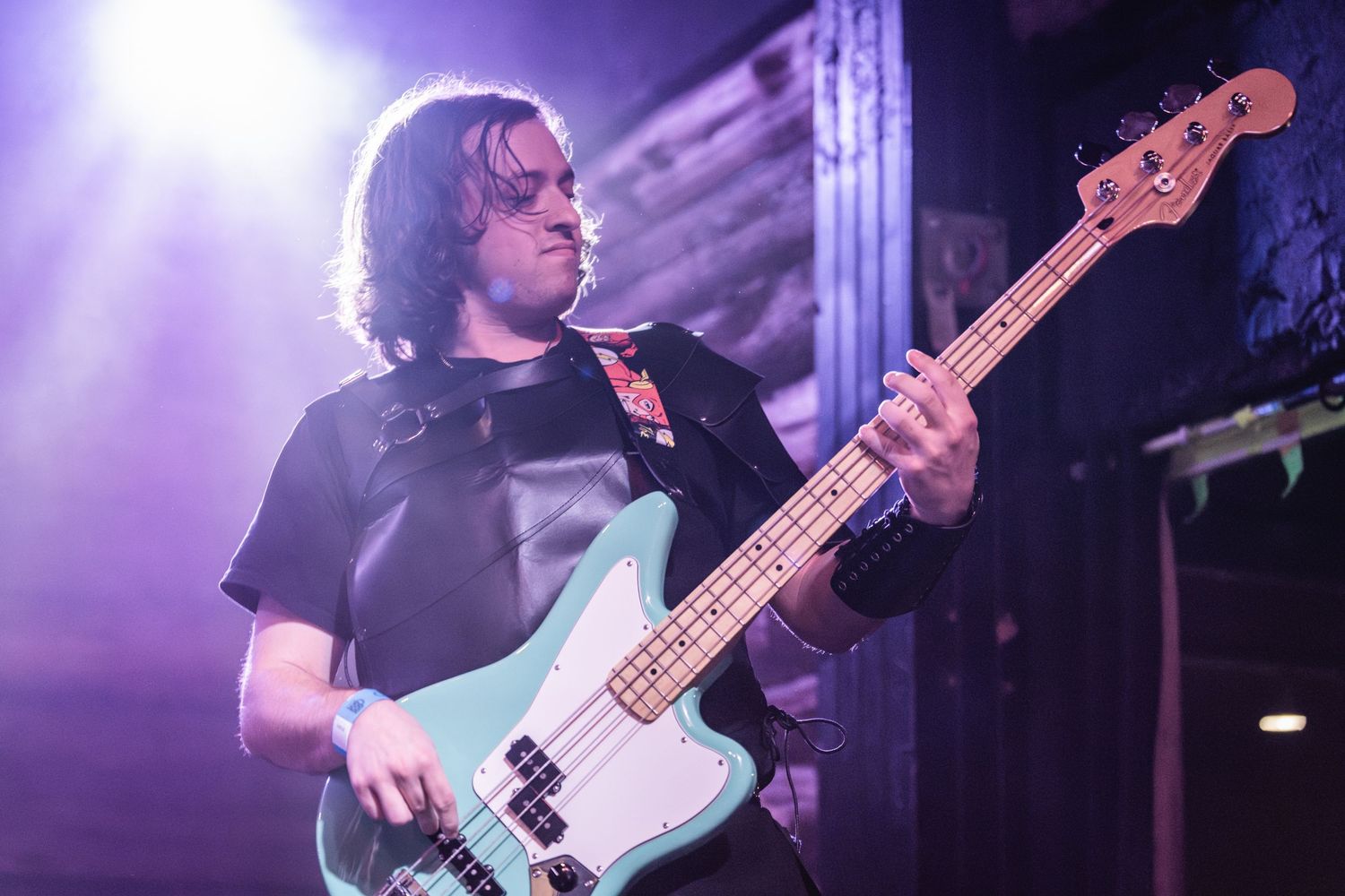 Bass guitarist performs on stage with purple lighting and smoke effects during a live concert performance.
