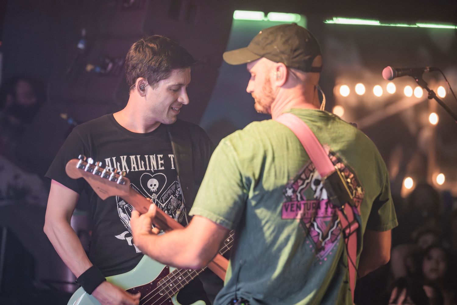 Two musicians perform together on stage with guitars under colorful stage lighting at a live concert performance.