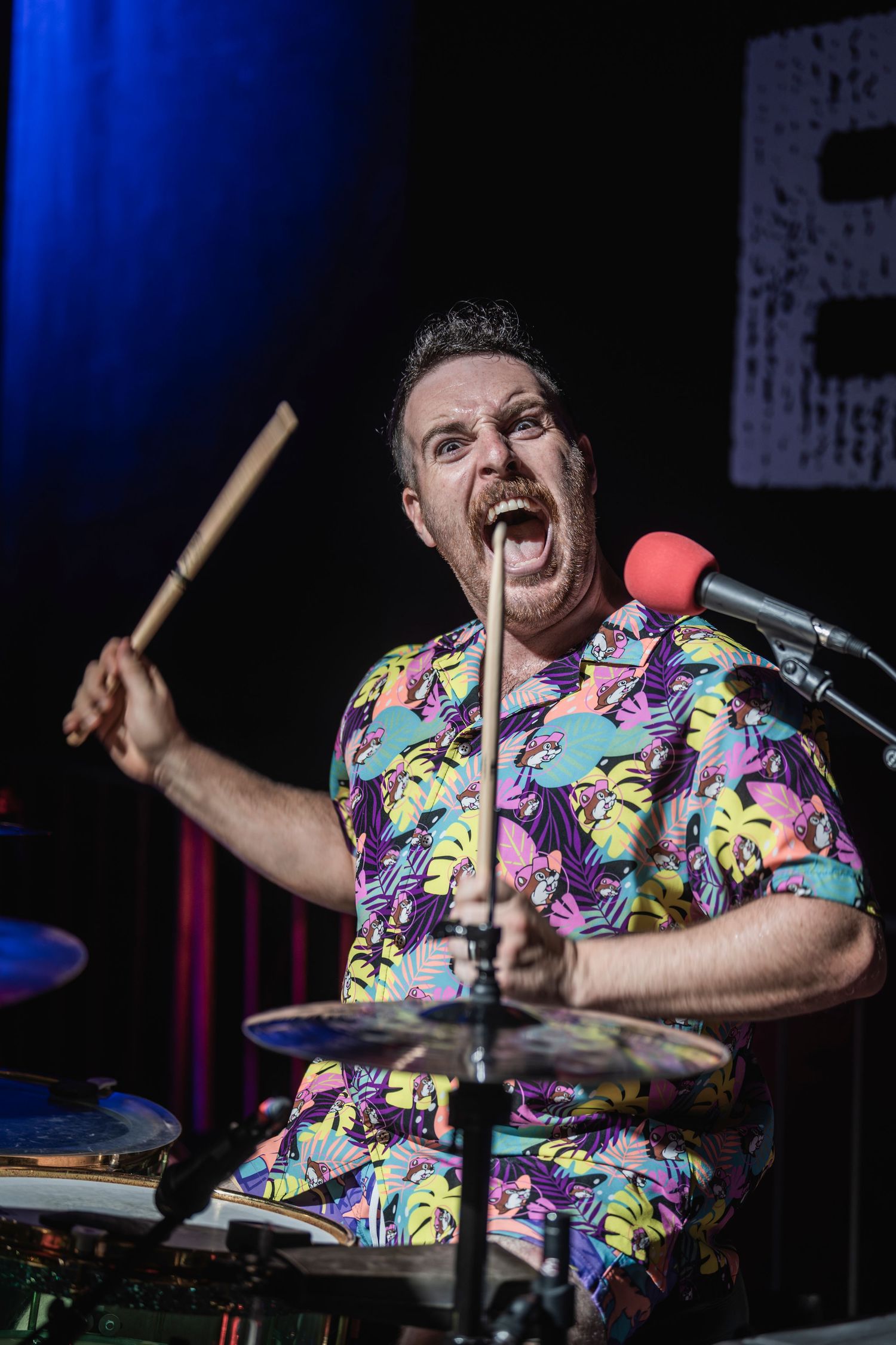 Energetic drummer in colorful patterned shirt performs passionately on stage with dramatic lighting.