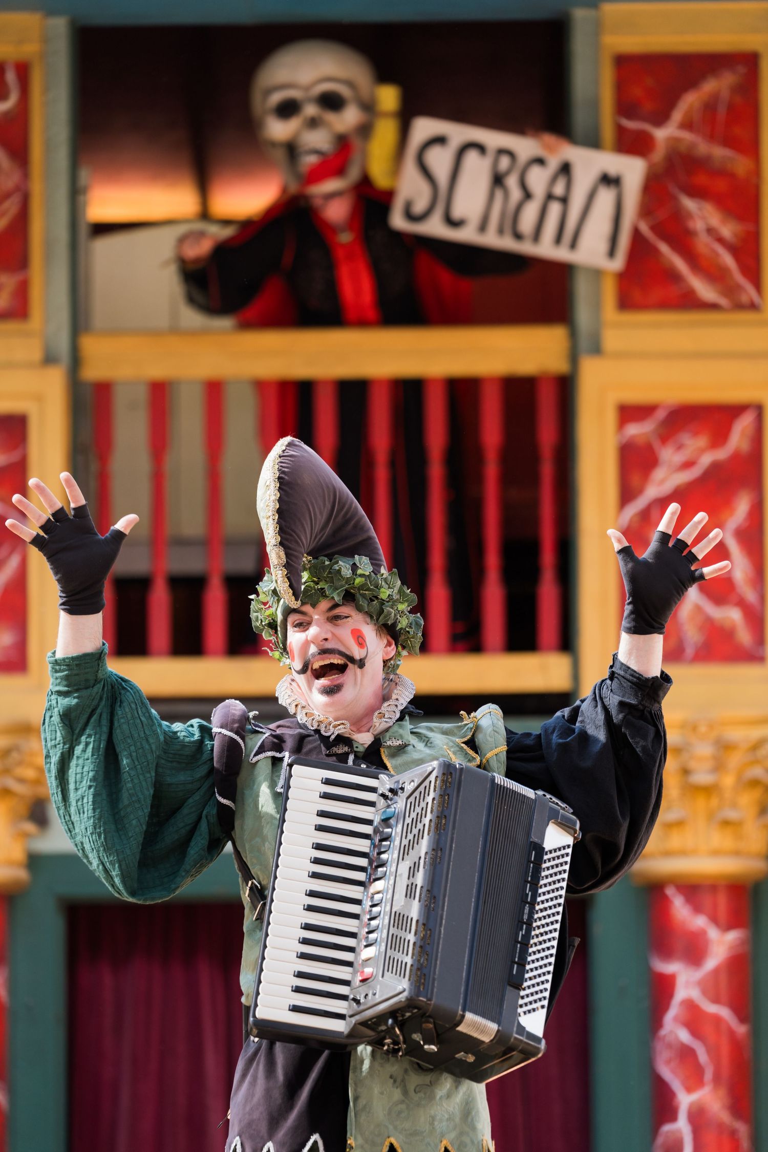 A performer in a theatrical costume plays an accordion with raised arms on stage with colorful backdrop and props.