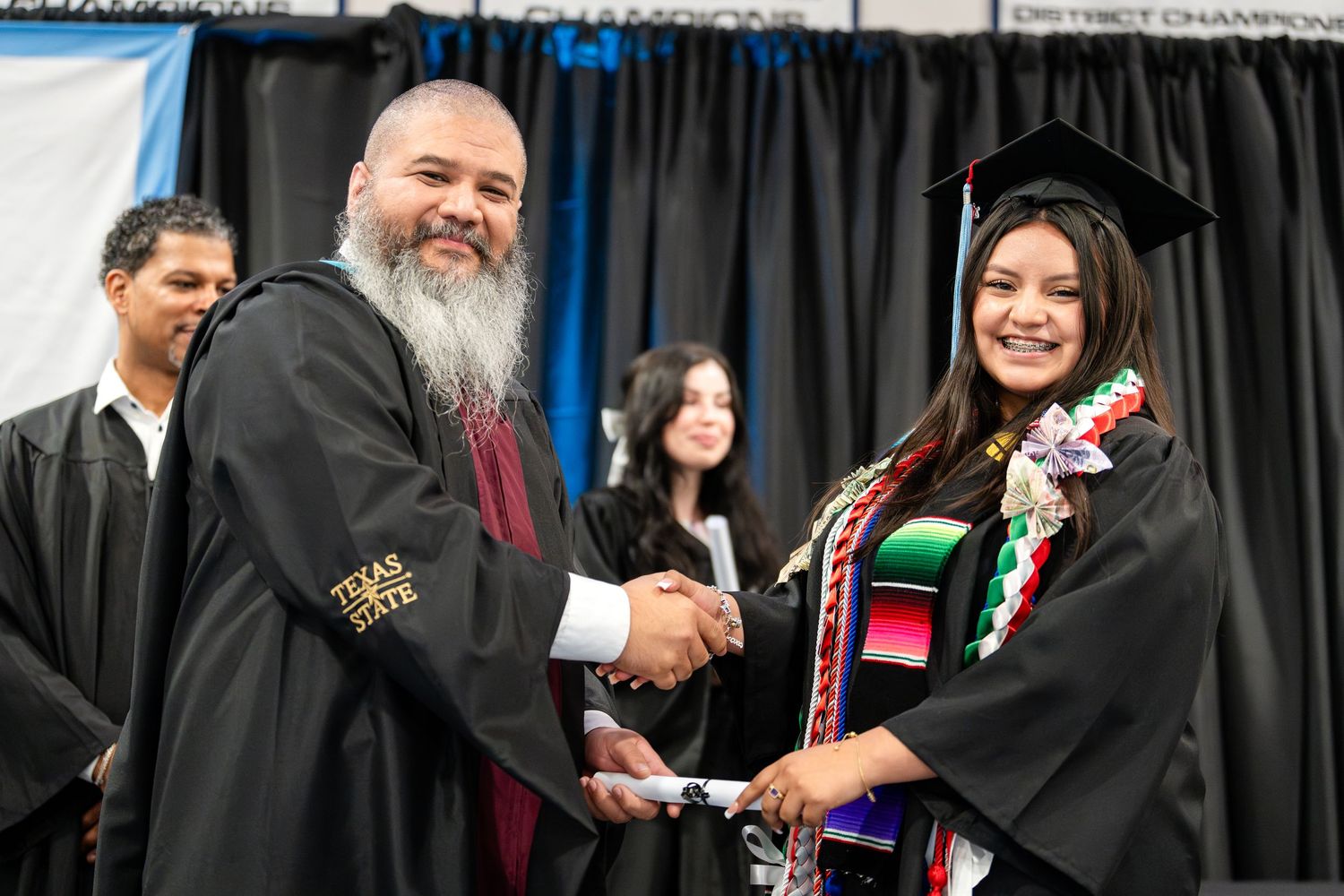 Graduates in black caps and gowns receiving diplomas during a commencement ceremony.