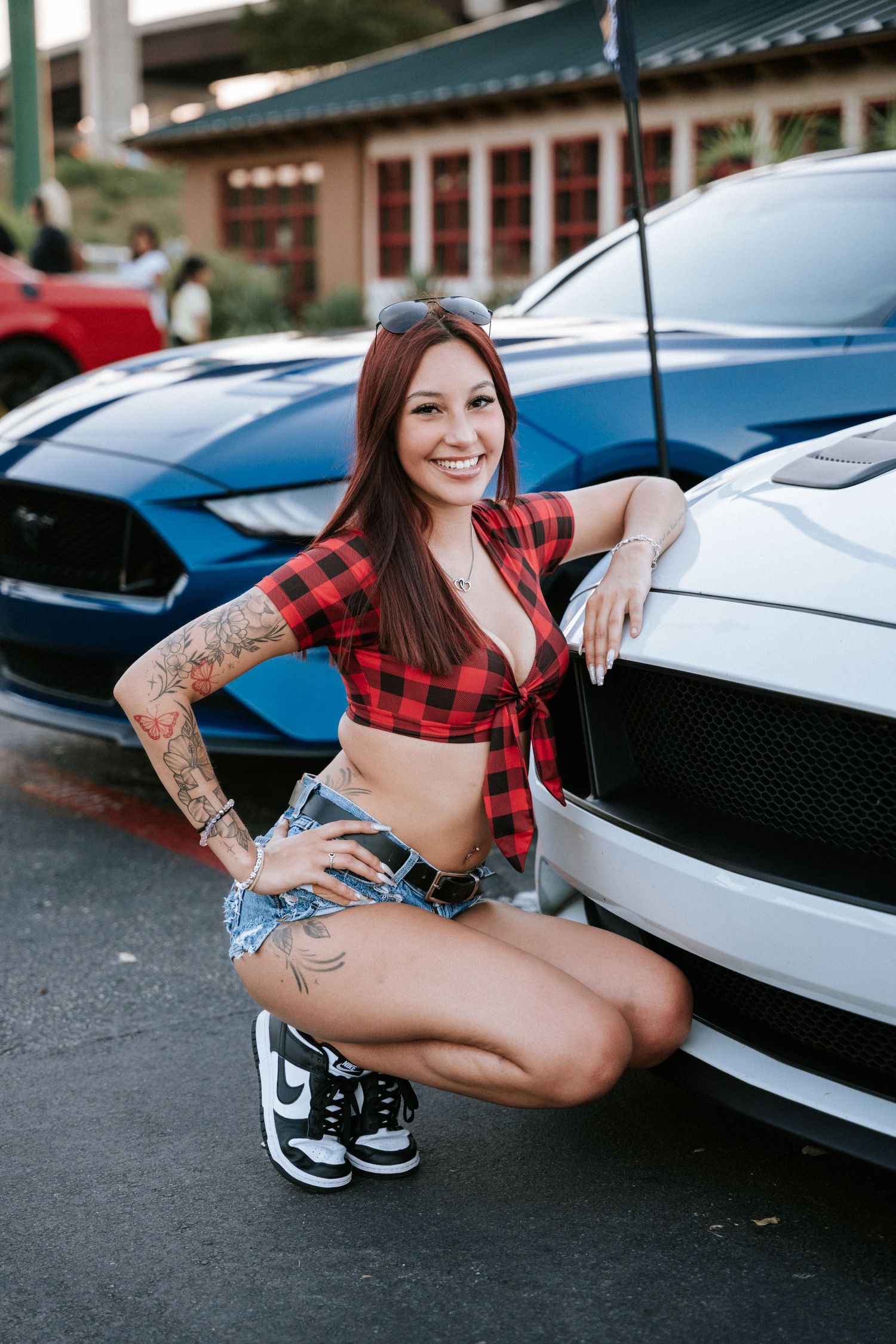 Model in red plaid crop top and denim shorts poses with white sports car in parking lot.
