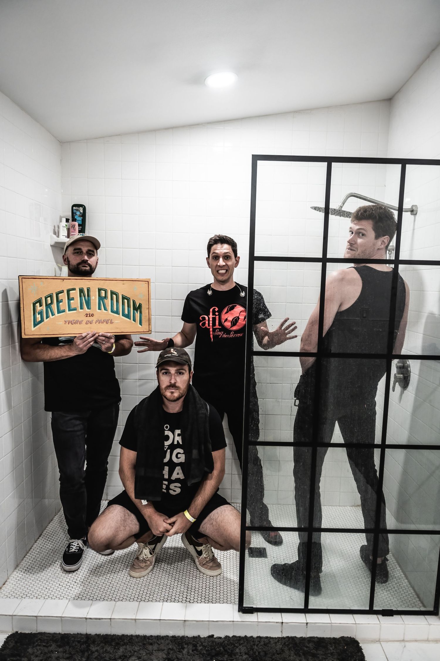 Musicians pose with a Green Room sign in a white tiled bathroom setting.