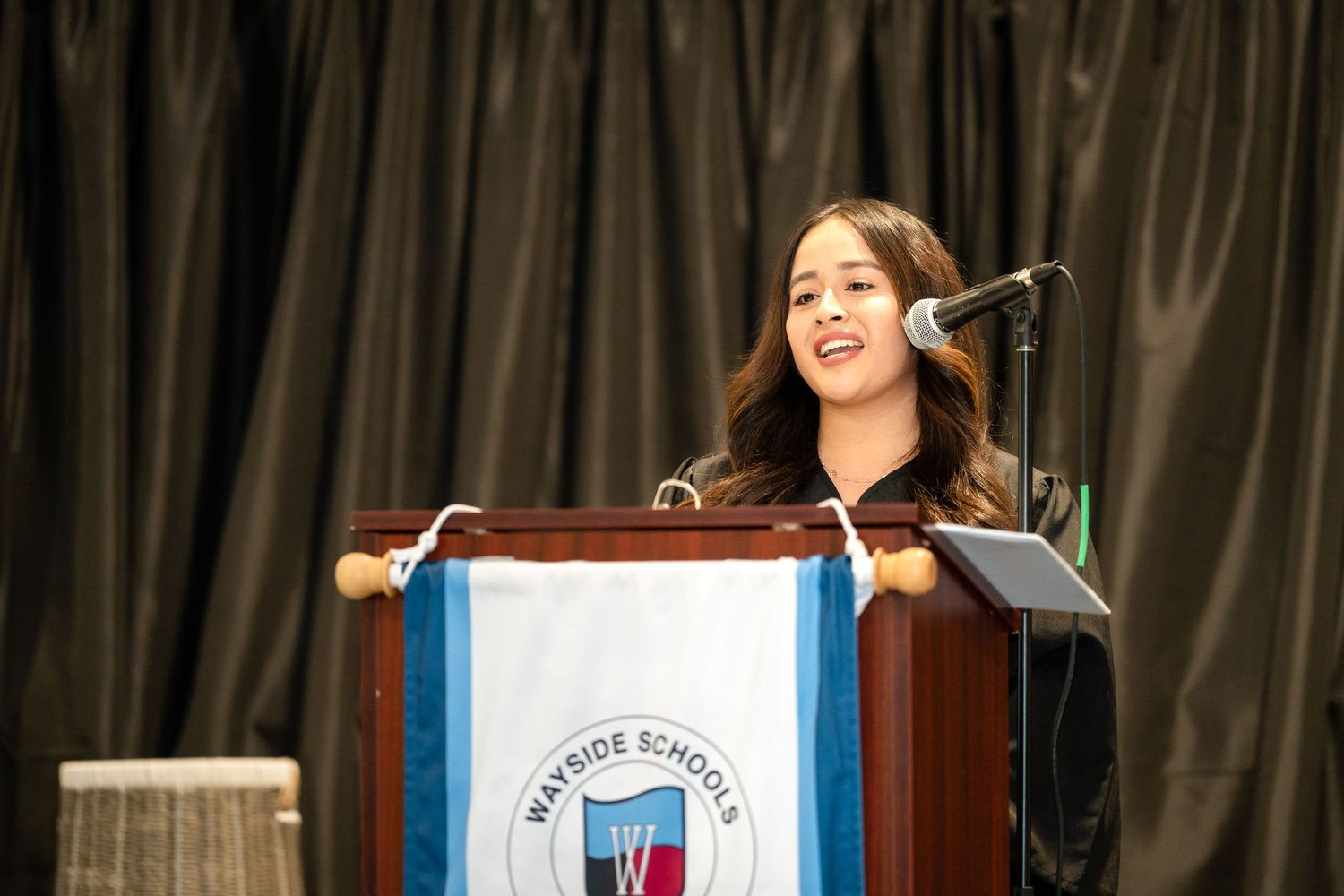 A speaker stands at a podium with a blue and white banner during a graduation ceremony.
