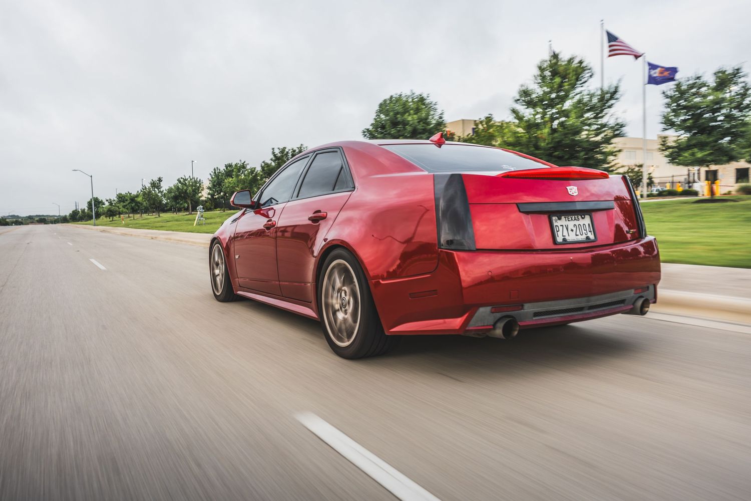 Red Cadillac CTS-V sedan driving on a suburban road with flags visible in background.