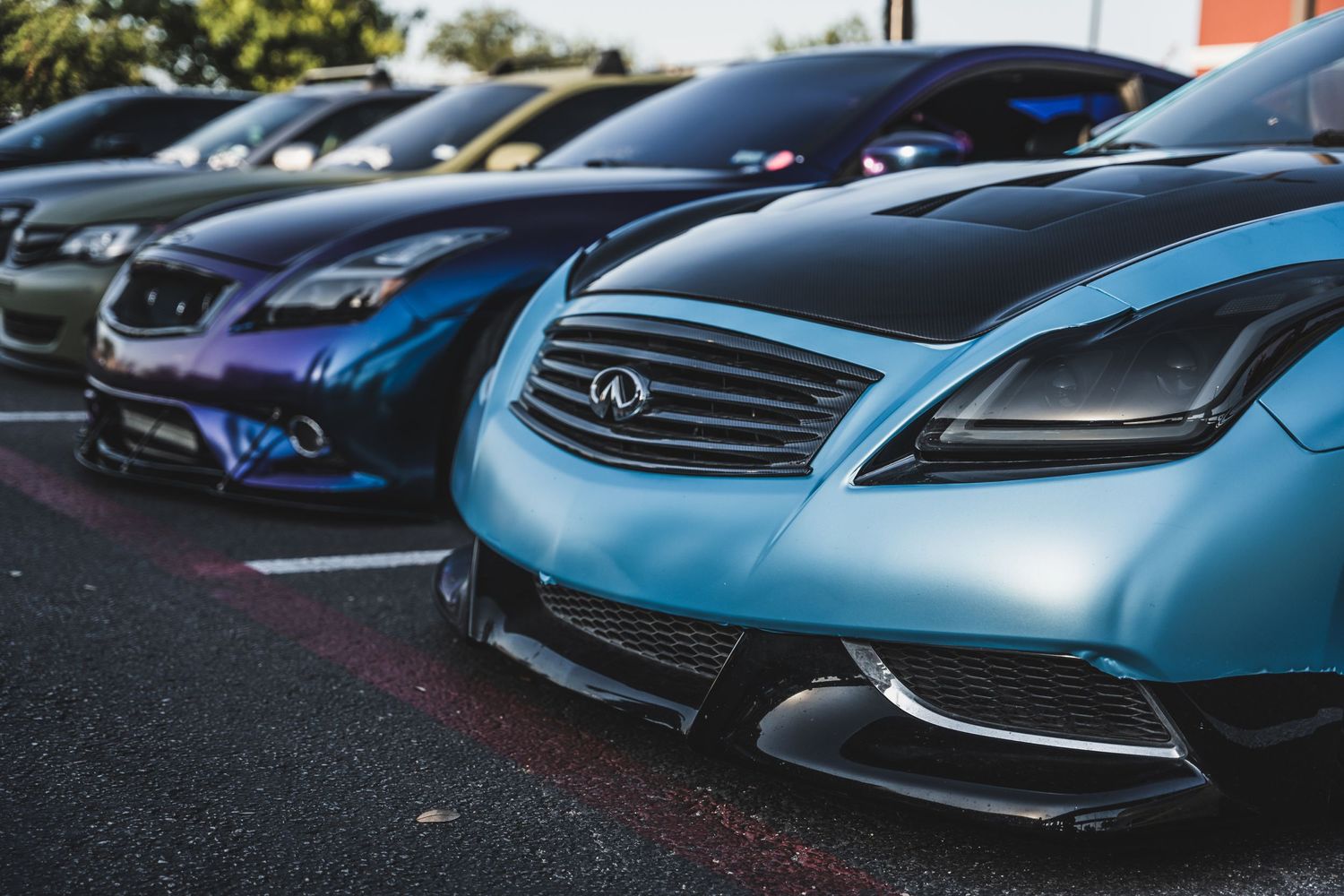 Row of luxury Infiniti cars in metallic colors parked at a car meet showing front grilles and headlights.