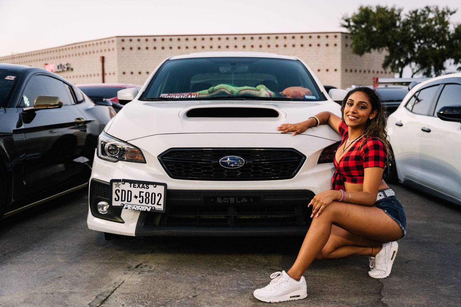 A person in red buffalo plaid crop top and jean shorts poses with a Subaru WRX in a parking lot.