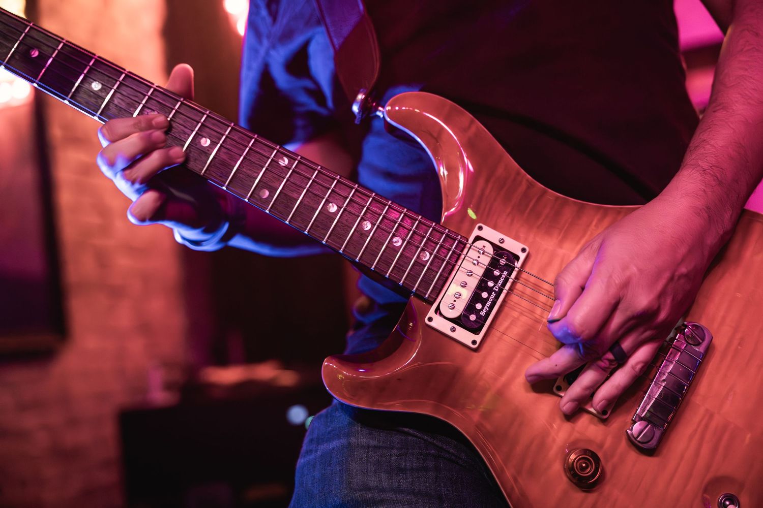Close-up shot of hands playing an electric guitar with wood grain finish under colored stage lights.
