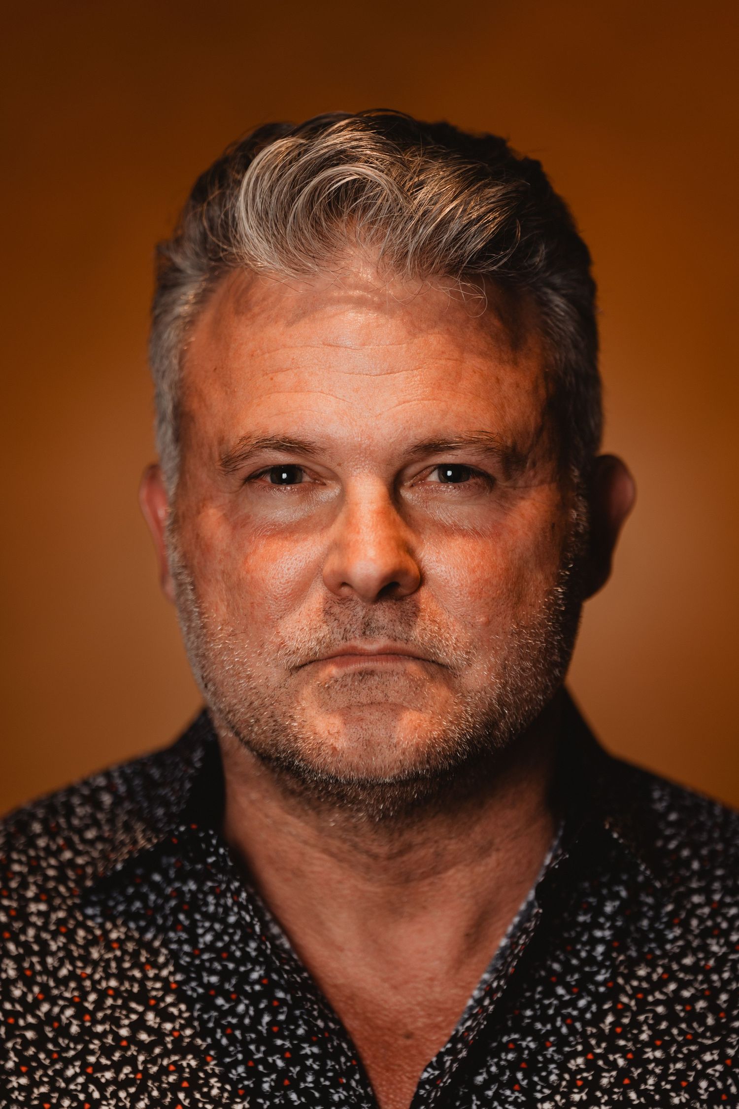 Close-up headshot of a man with salt and pepper hair wearing a patterned dark shirt against an orange backdrop.