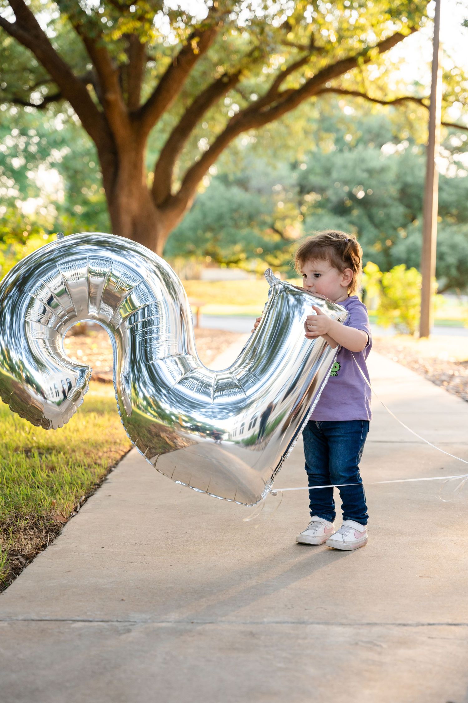 A toddler in purple shirt and jeans plays with a large silver number balloon outdoors.