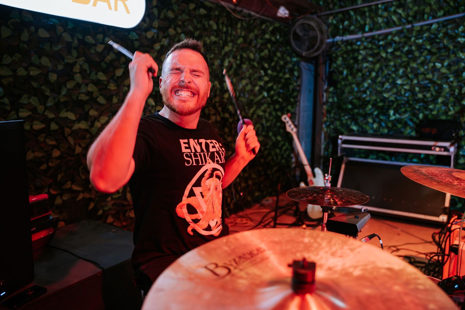 Drummer performing passionately on stage under red lighting at a concert venue.