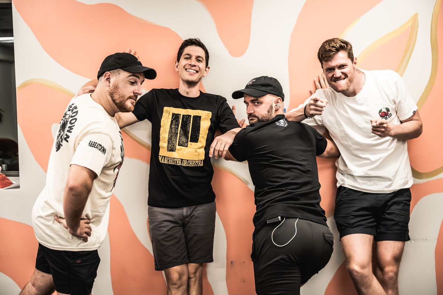 Four performers pose playfully against a peach-colored wall with white swirl patterns.
