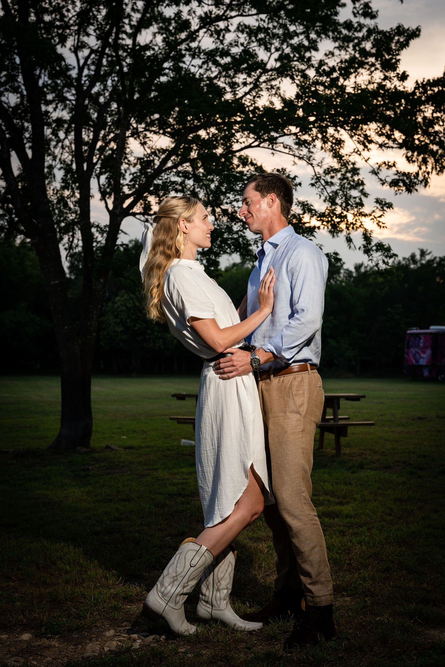 A couple embraces in a park at sunset, with her wearing stylish white boots.