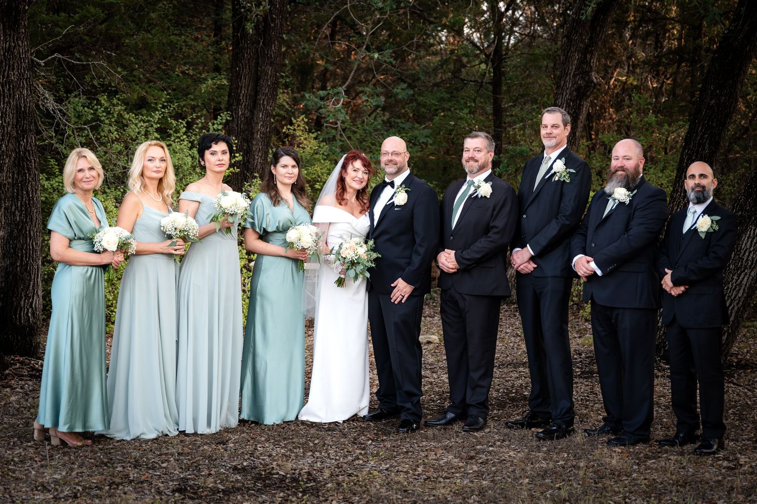 Wedding party poses outdoors with bridesmaids in blue dresses and groomsmen in dark suits.