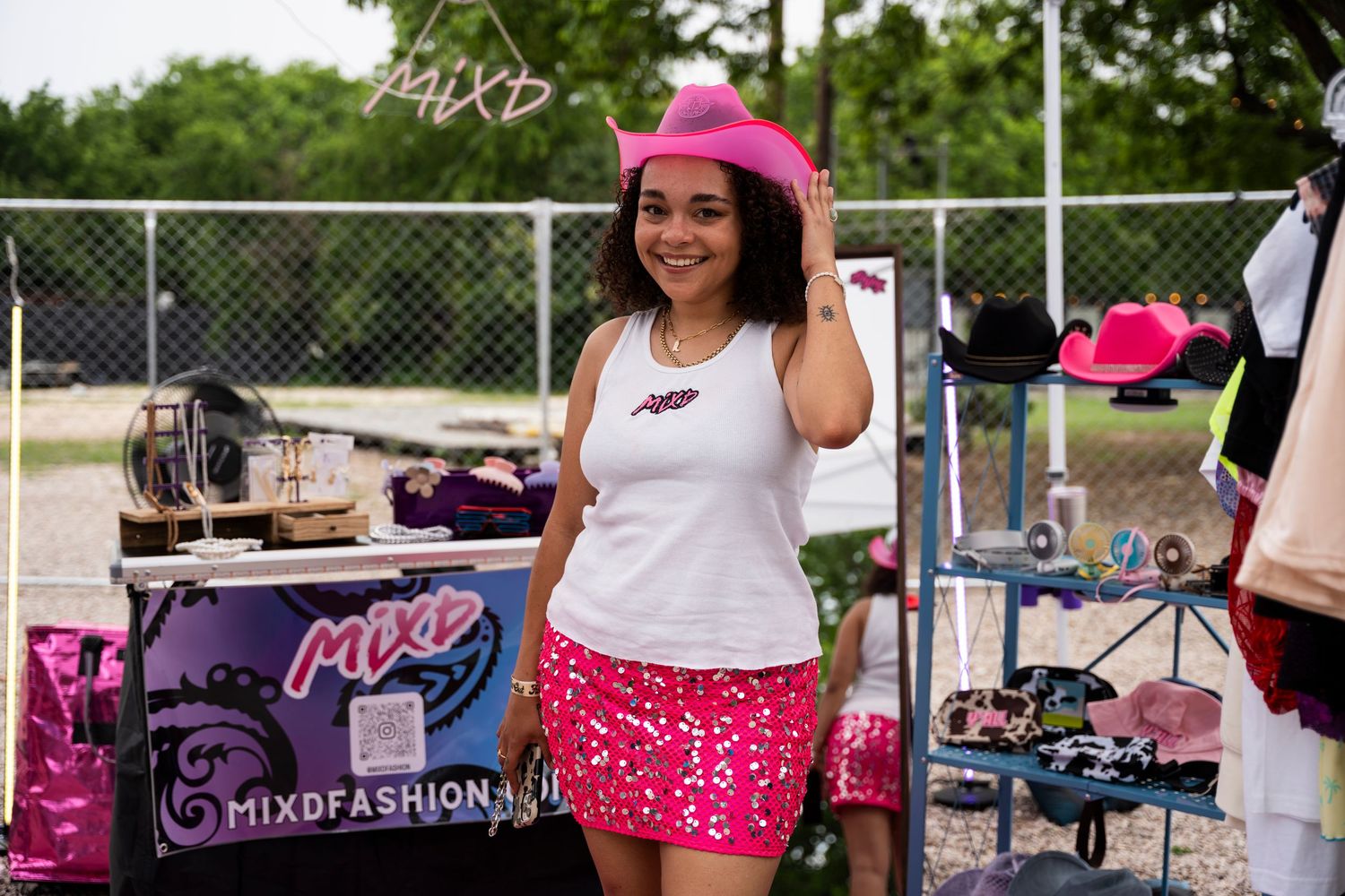 A cheerful vendor in a pink baseball cap and matching skirt stands behind their outdoor fashion display table.