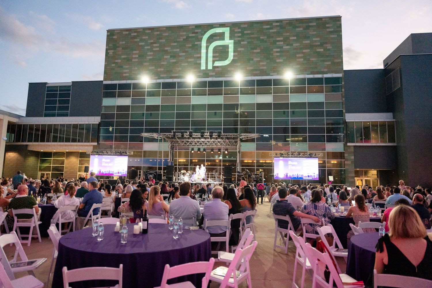 Evening event space with illuminated building facade and tables arranged on outdoor plaza.