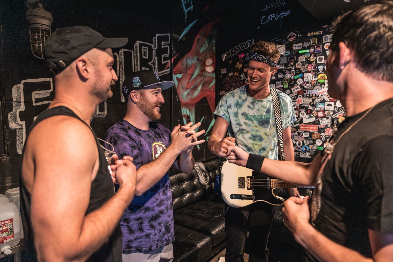 Musicians interact backstage against a wall covered in stickers and posters.