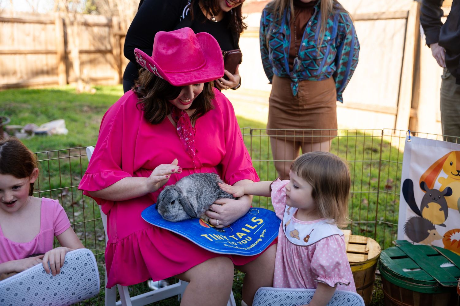 A group gathers in a backyard garden while a person in a bright pink dress holds a small grey kitten.