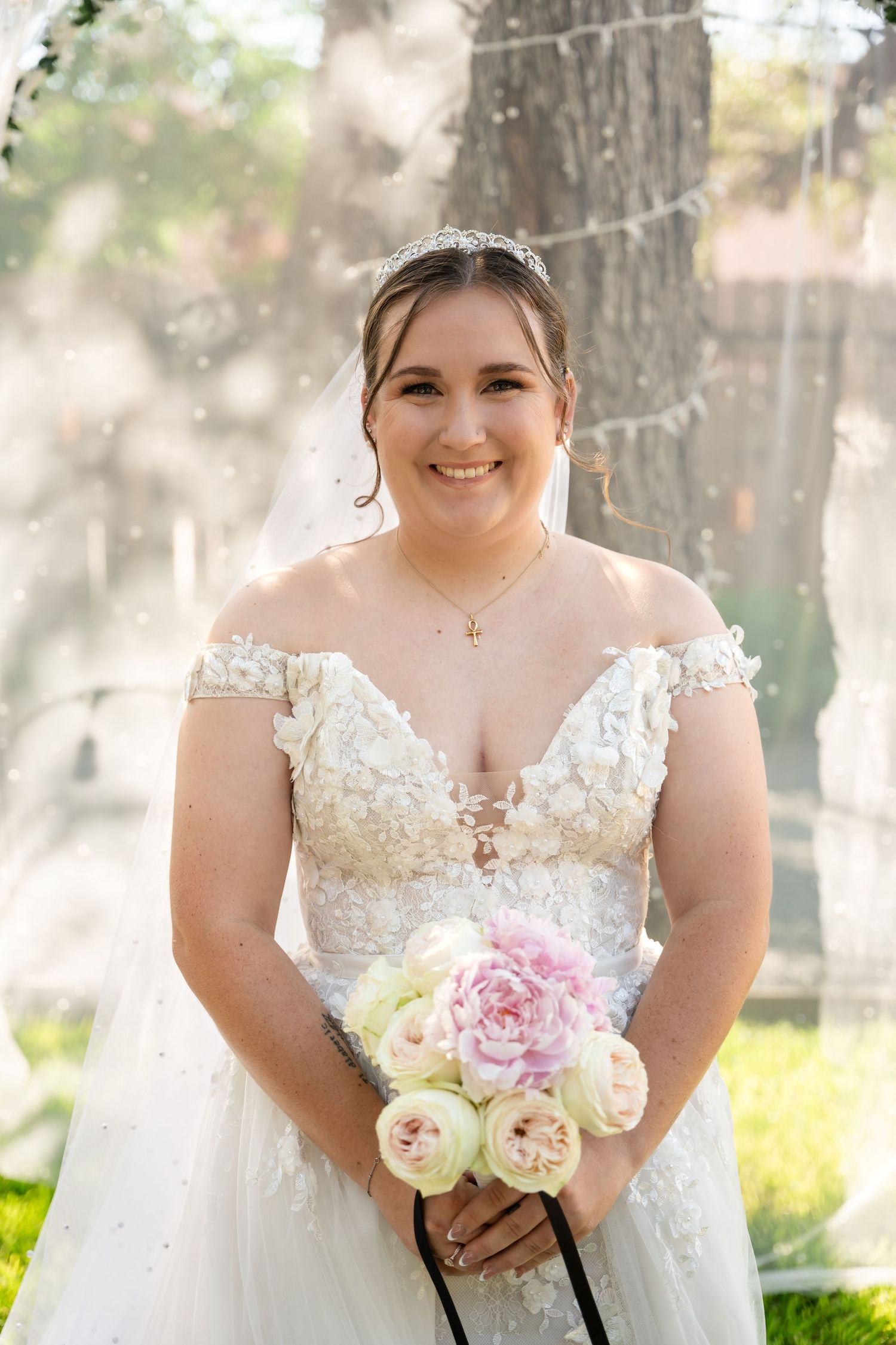 A bride holds a bouquet of pink and white peonies in a sunlit outdoor setting.