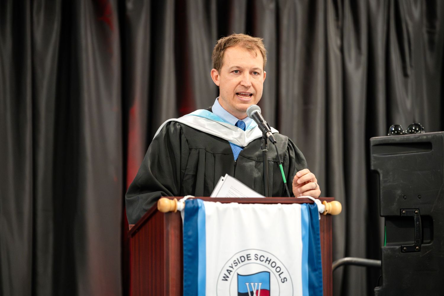 Faculty member in academic regalia speaks at podium during graduation ceremony.