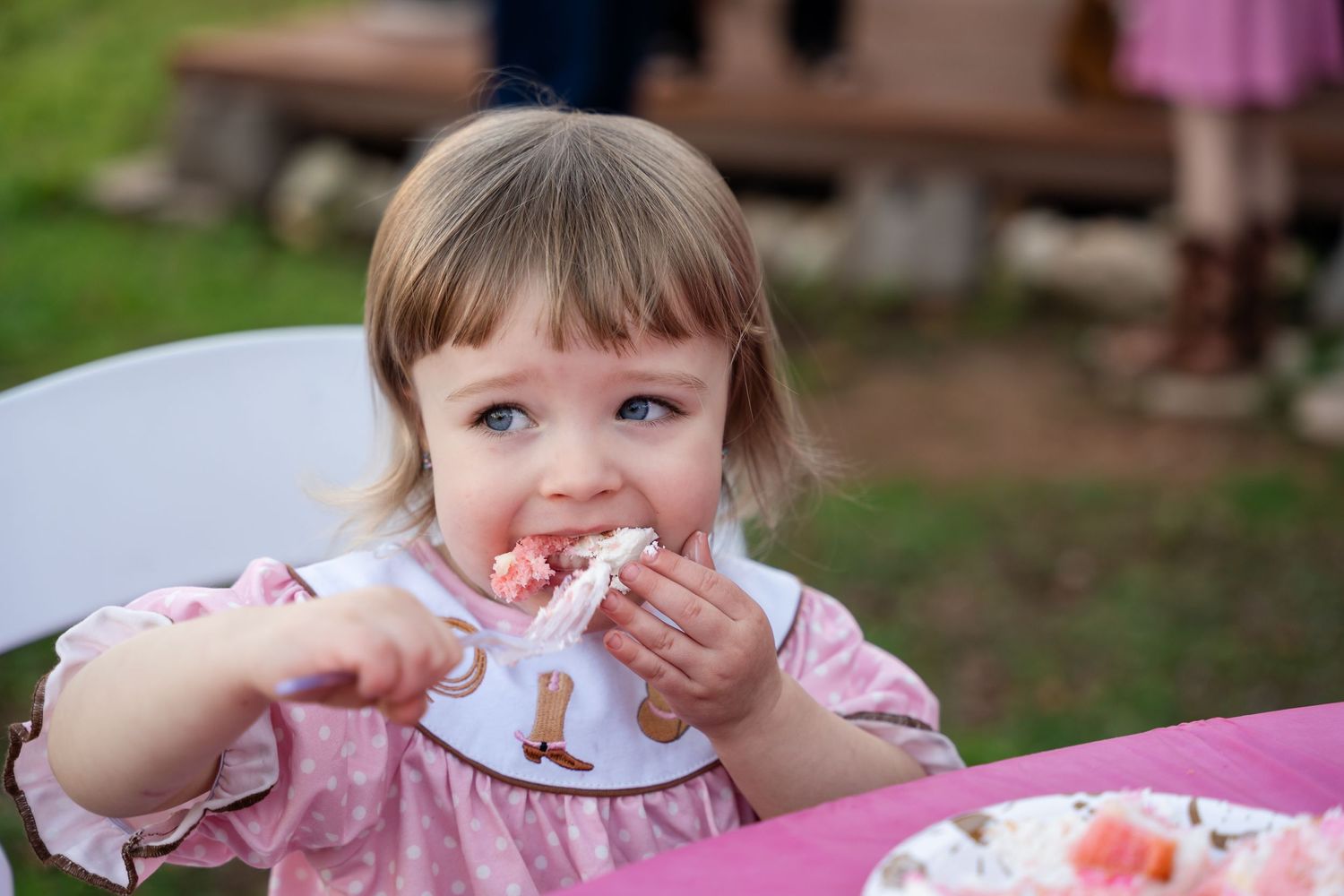 Young child in pink shirt sitting at outdoor table eating and making playful expression during meal time.