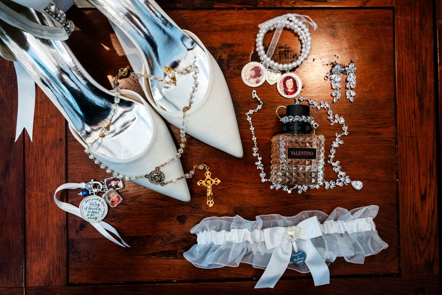 Wedding accessories including white heels, jewelry, and a garter laid out on dark wooden surface.