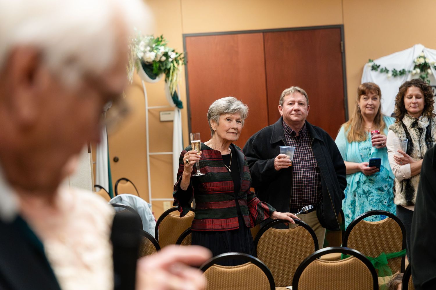 A group of people holding drinks and mingling in a room with padded chairs at an informal gathering.