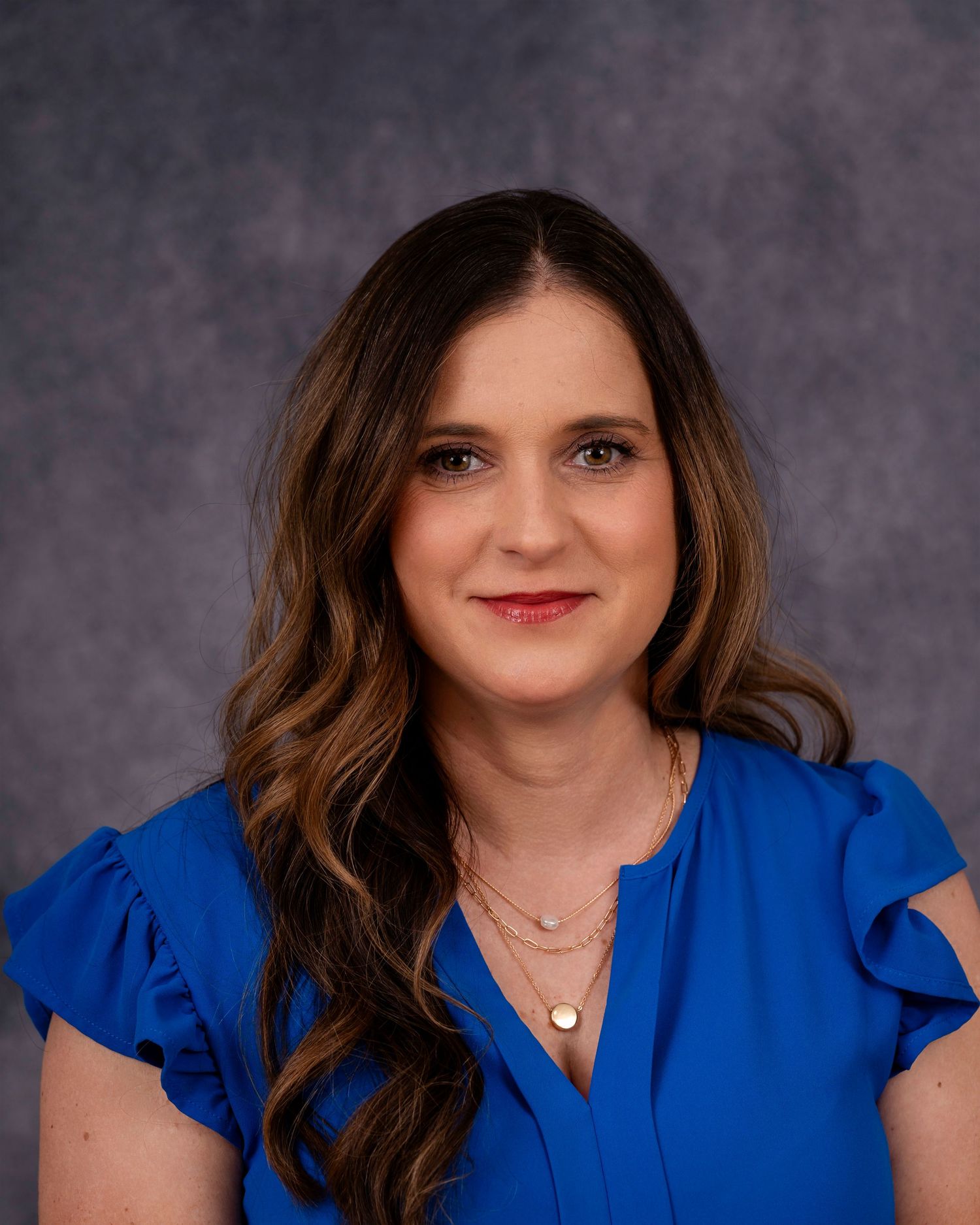 Professional headshot portrait of a person in a royal blue blouse with ruffled sleeves against a gray background.