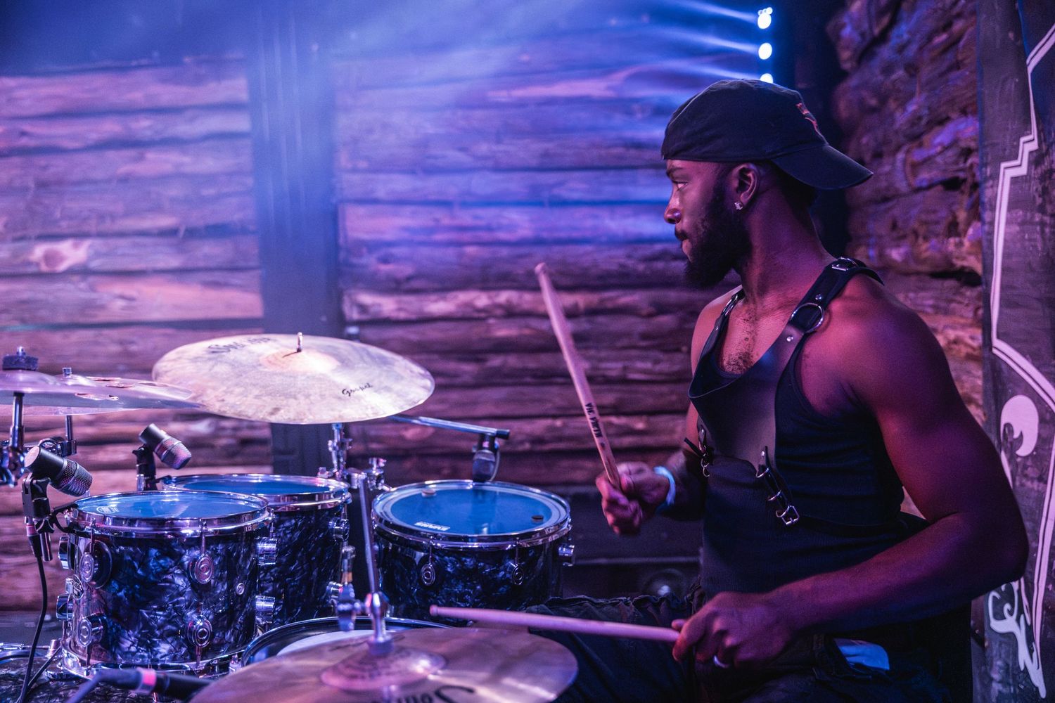 Drummer plays with intensity on stage with dramatic lighting and wooden wall backdrop.