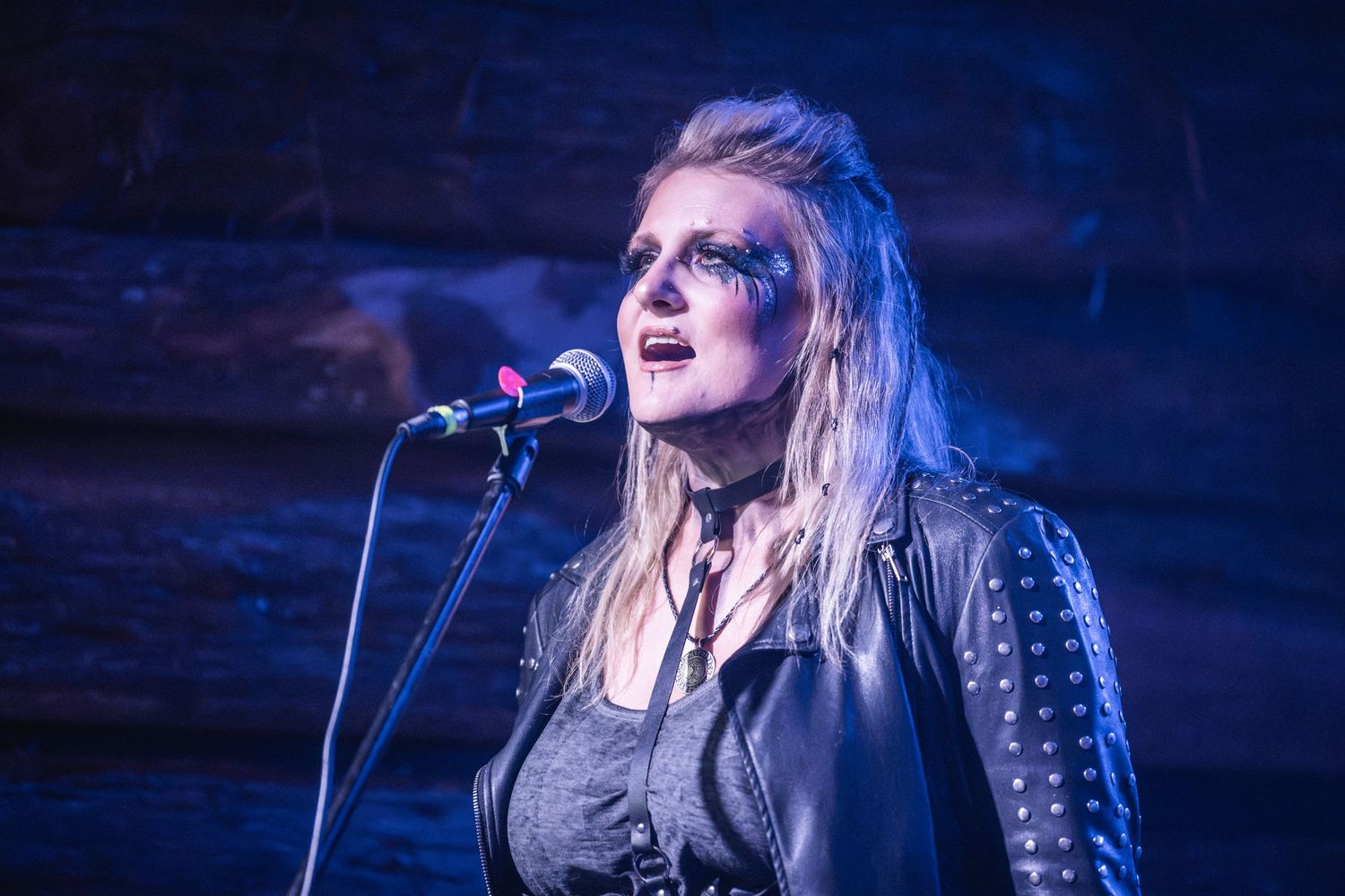 Dramatic close-up of performer with stage makeup singing into microphone under blue lights.