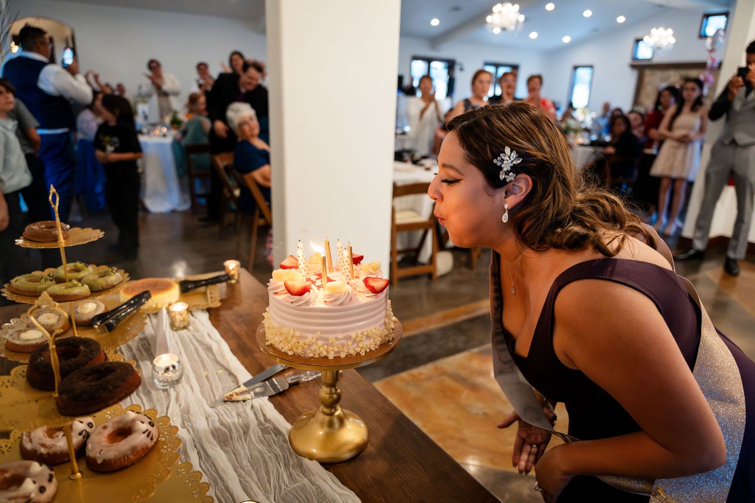Someone in a black dress blows out birthday candles on a cake while party guests watch in the background.