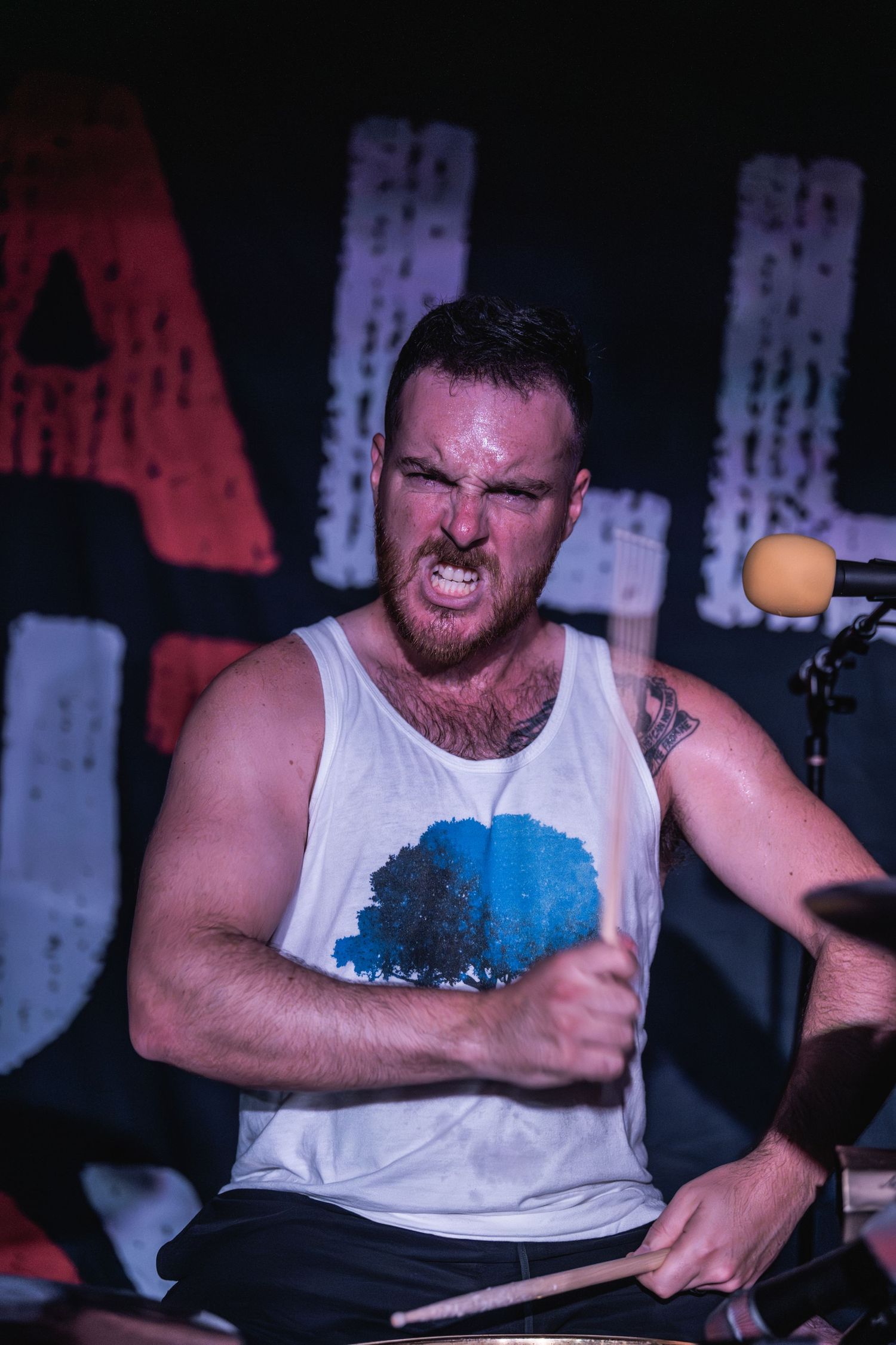 Intense drummer performs with passionate expression in white tank top on concert stage.