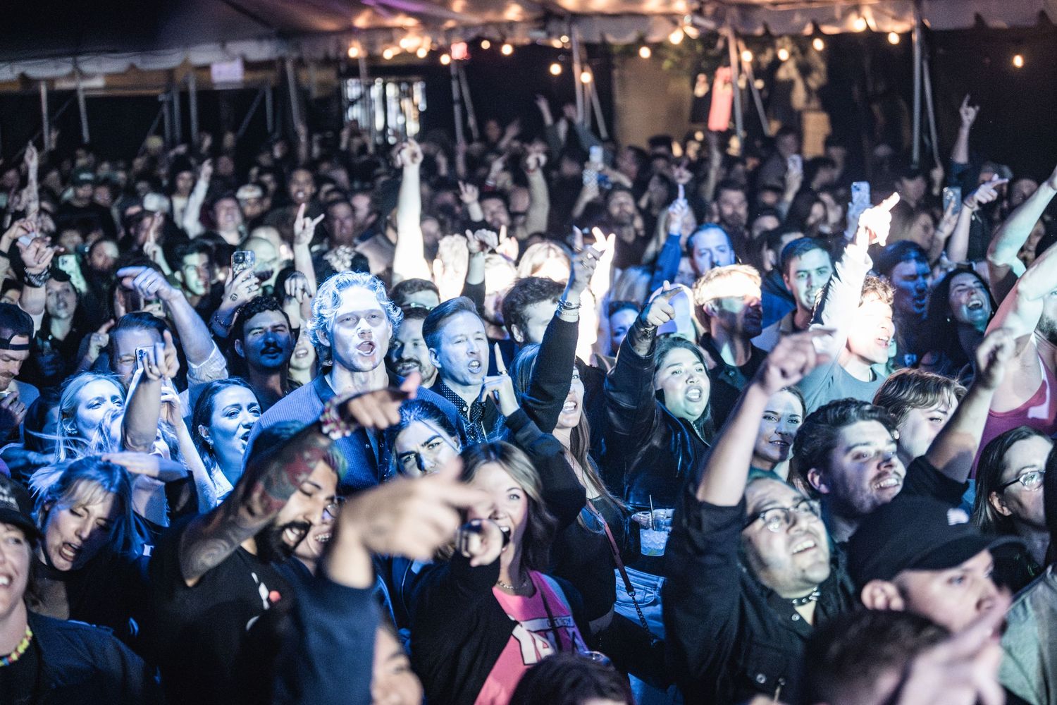 An energetic concert crowd illuminated by blue stage lights raises their hands during a live music performance.