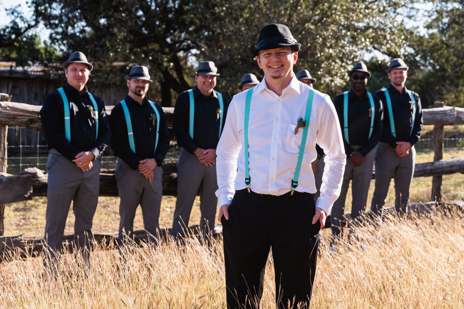Groomsmen in matching black suits and turquoise suspenders pose in a sunlit field.
